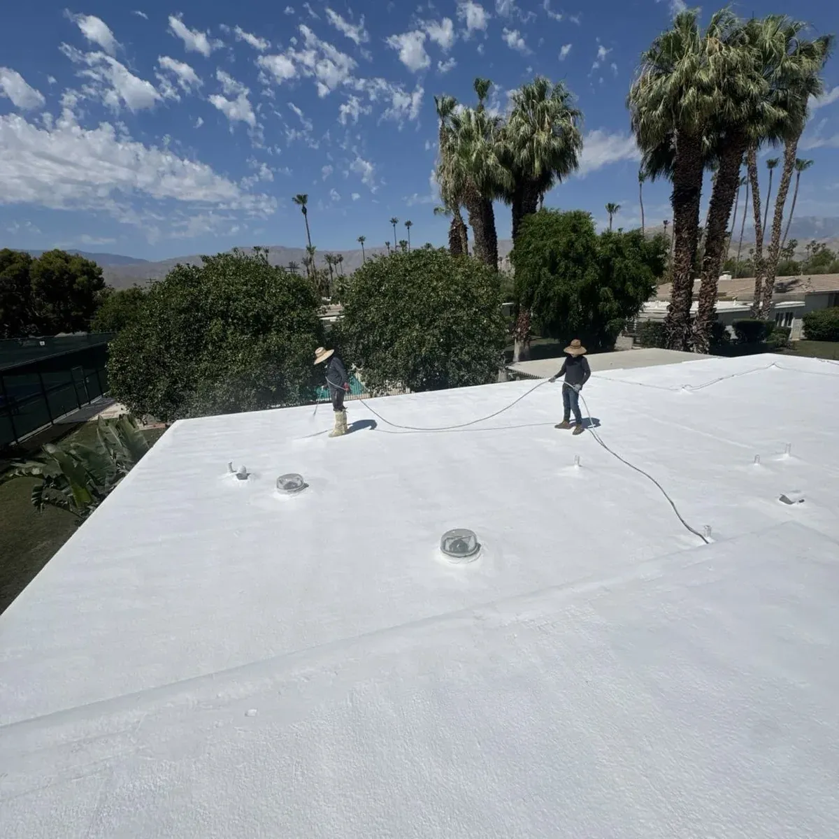 Two roofers finishing a freshly coated white SPF roof on a valley home, palms and mountains behind