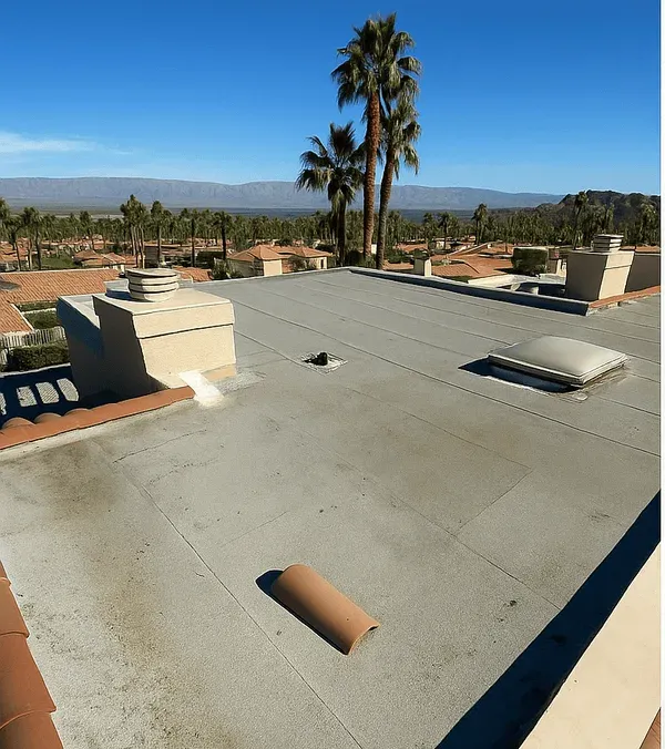 A low-slope roof before renewal: a weathered grey deck with staining and a dislodged tile, terracotta parapet edging, valley rooftops beyond